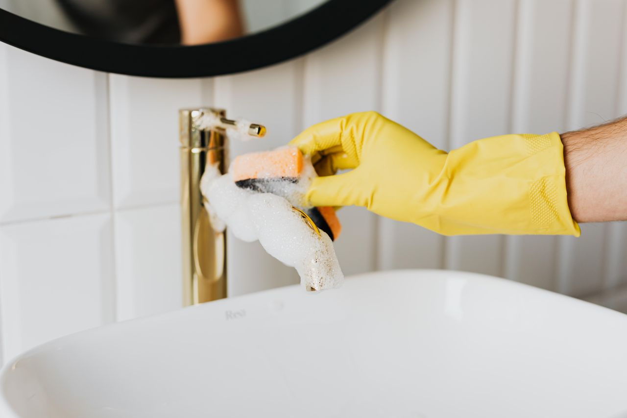 Un homme méconnaissable portant des gants en latex jaunes nettoie un robinet doré dans la salle de bain tout en faisant le ménage à la maison avec une éponge orange et du détergent